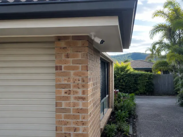 Exterior CCTV camera mounted under the eave on a brick home beside a driveway in Nerang